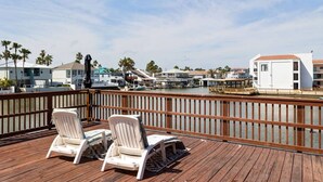 Una terraza de madera con dos sillones blancos, vista a un puerto deportivo con barcos y edificios al fondo.
