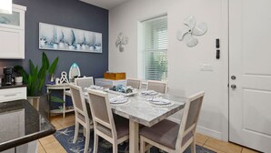 Un comedor con una mesa dispuesta para cuatro, una cocina con cafetera y un cuadro grande en la pared.