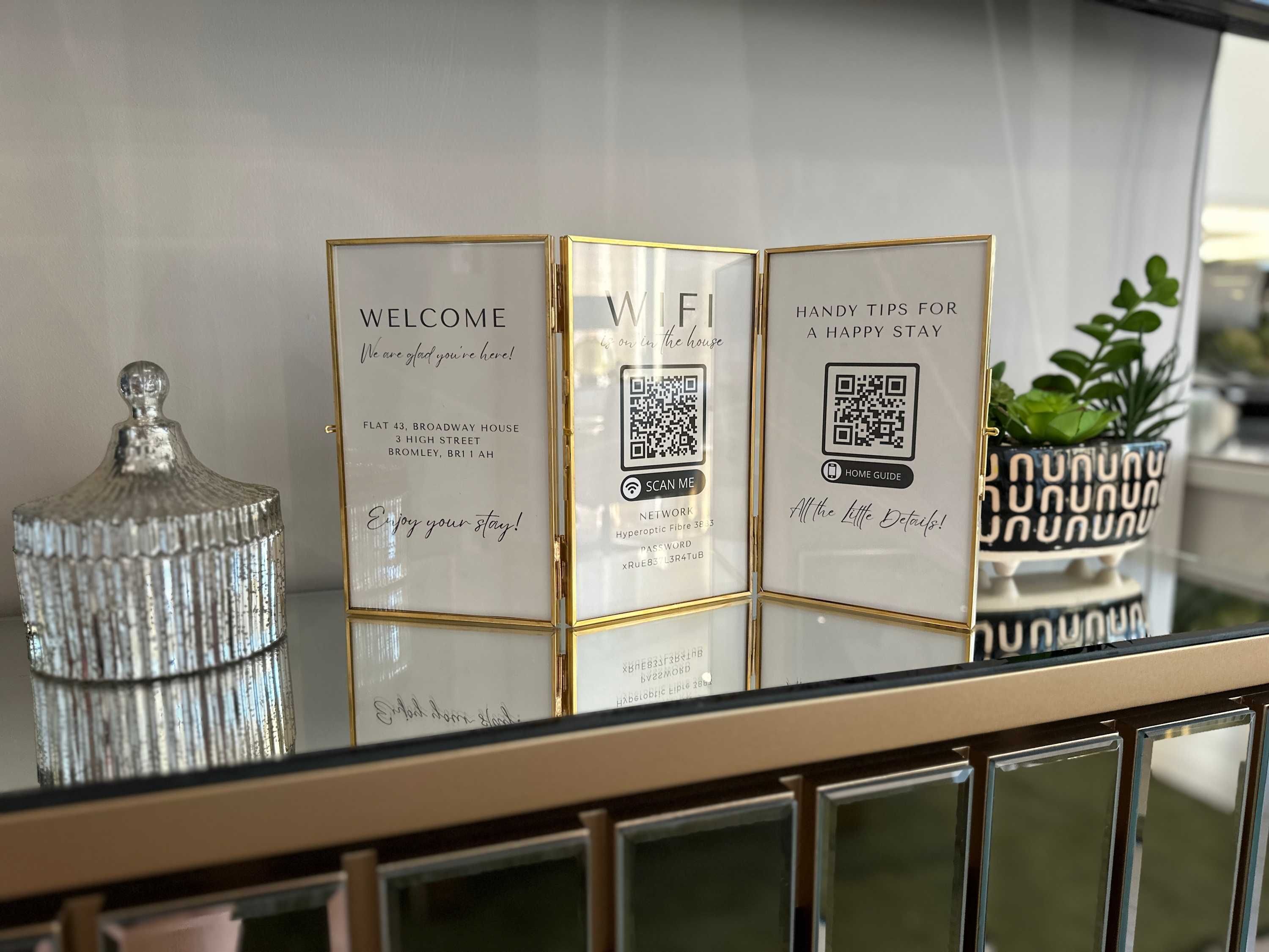 Trois panneaux d’accueil encadrés, chacun muni d’un code QR, un bocal décoratif en verre et une plante en pot, le tout posé sur une surface réfléchissante.