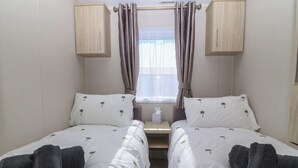 A twin room with two beds, each with a pillow and a towel, a window with curtains, and wooden cabinets. - Pheasant Lodge (Holyhead)