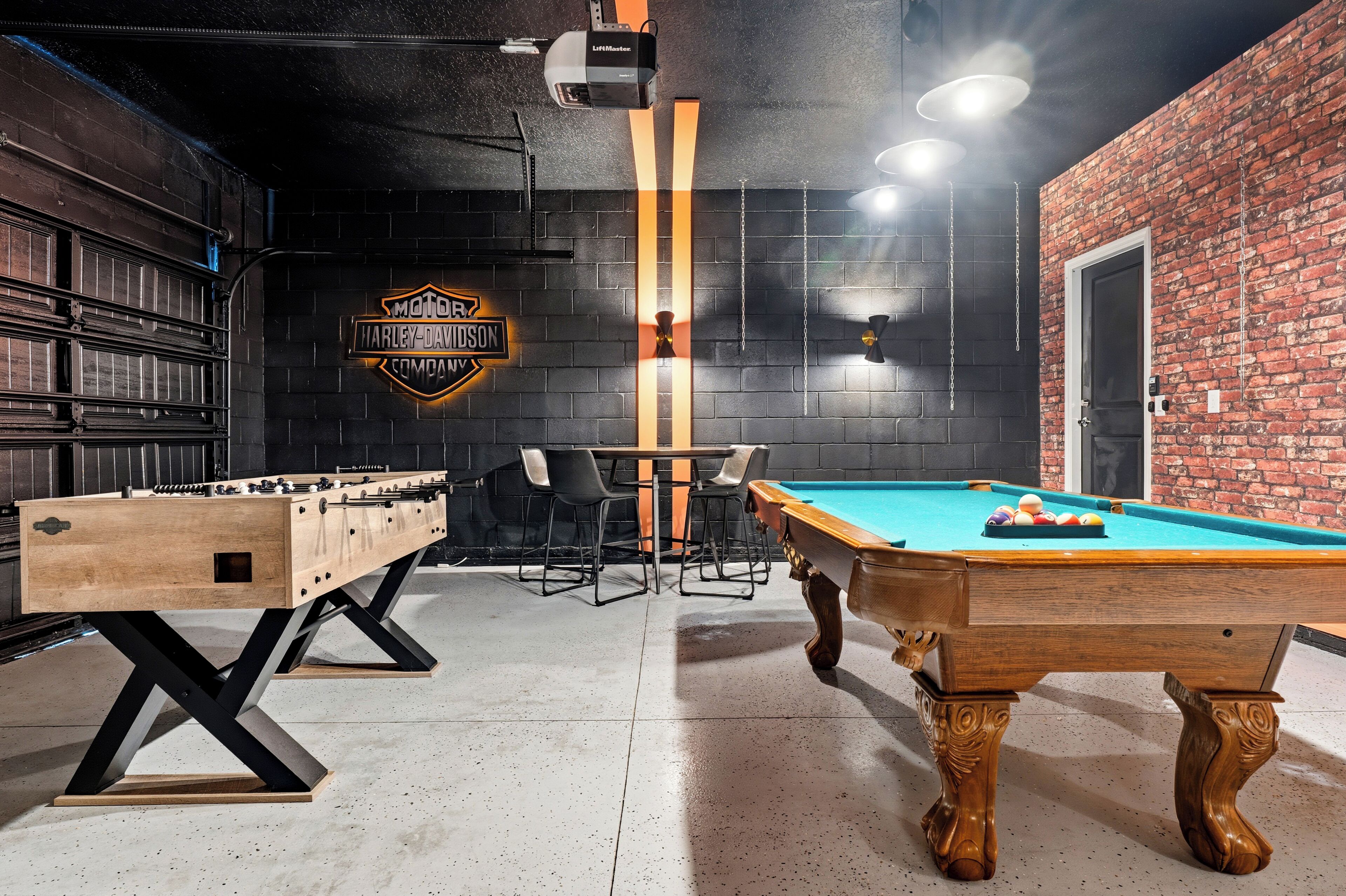 Uma mesa de bilhar, uma mesa de futebol de mesa e um letreiro Harley-Davidson em um cômodo com paredes de tijolo e uma porta.
