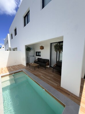 Uma área de estar externa moderna com piscina, piso de madeira, um sofá e uma mesa de jantar.