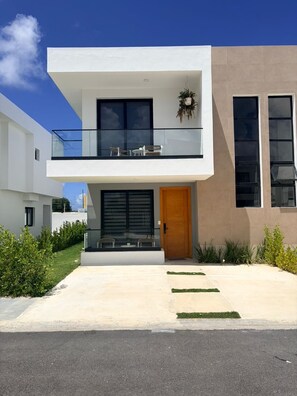 Ein modernes, zweistöckiges Haus mit Balkon, Eingangstür und einer kleinen Sitzgelegenheit im Freien.