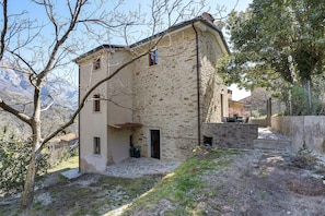 Un bâtiment en pierre avec un escalier en pierre menant à une porte, entouré d’arbres et avec une chaîne de montagnes en arrière-plan.