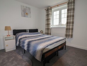 A bedroom with a bed, nightstand, lamp, window with curtains, radiator, and a wall-mounted flower painting. - Apartment 7 at Anfield (Liverpool)