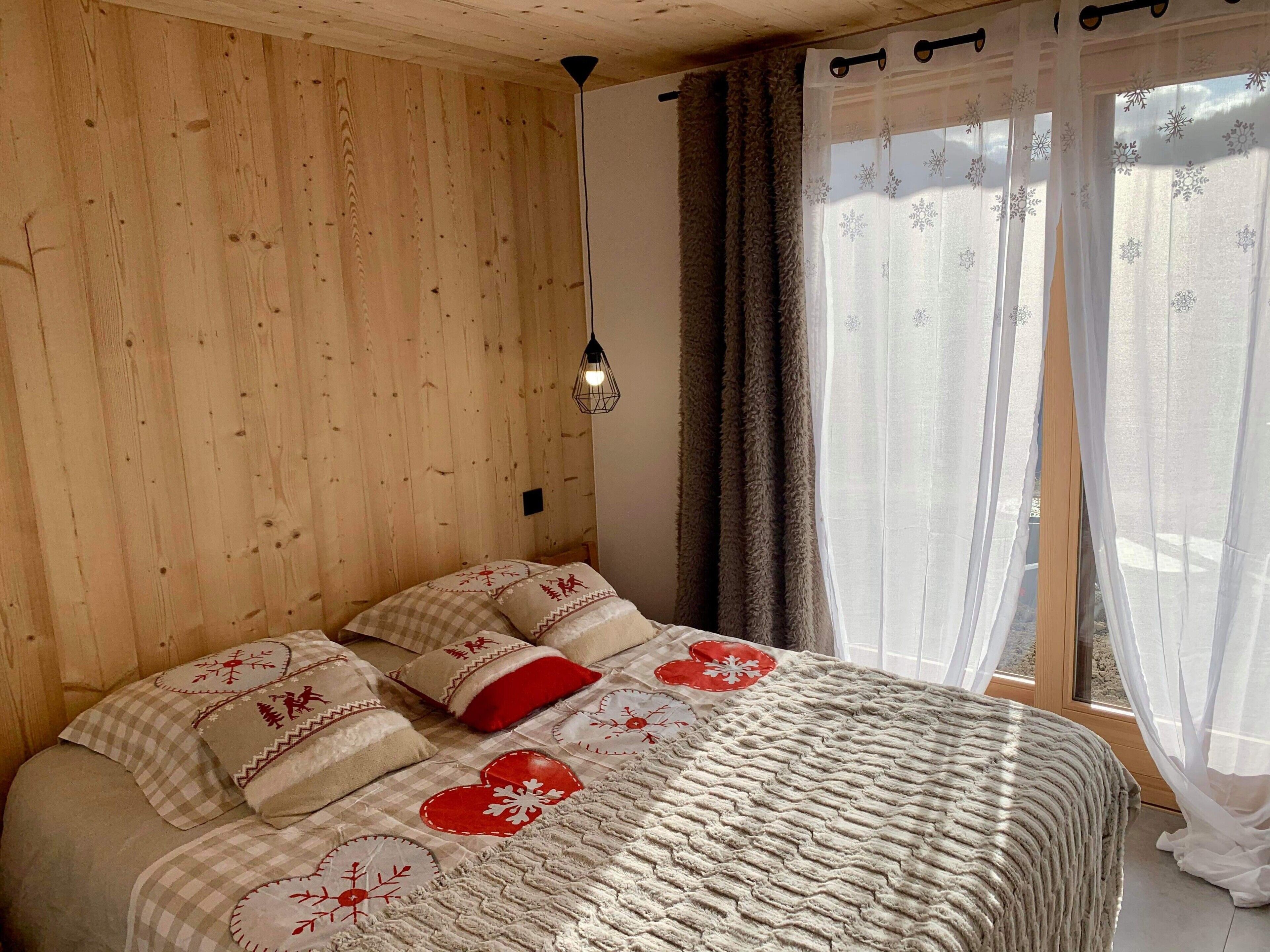 A wooden-paneled room with a bed, a hanging light fixture, and a window with sheer curtains.
