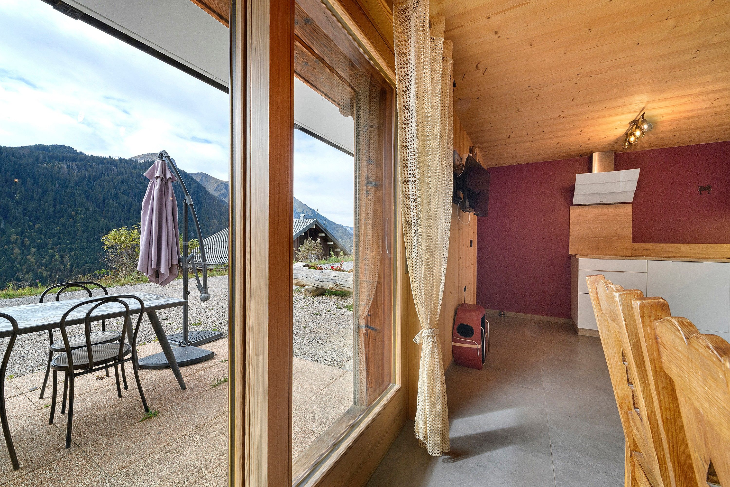 Ein Zimmer mit großem Fenster, Holzboden, einer Essecke mit Stühlen und Tisch sowie einer Küchenzeile mit Spüle und Schränken.