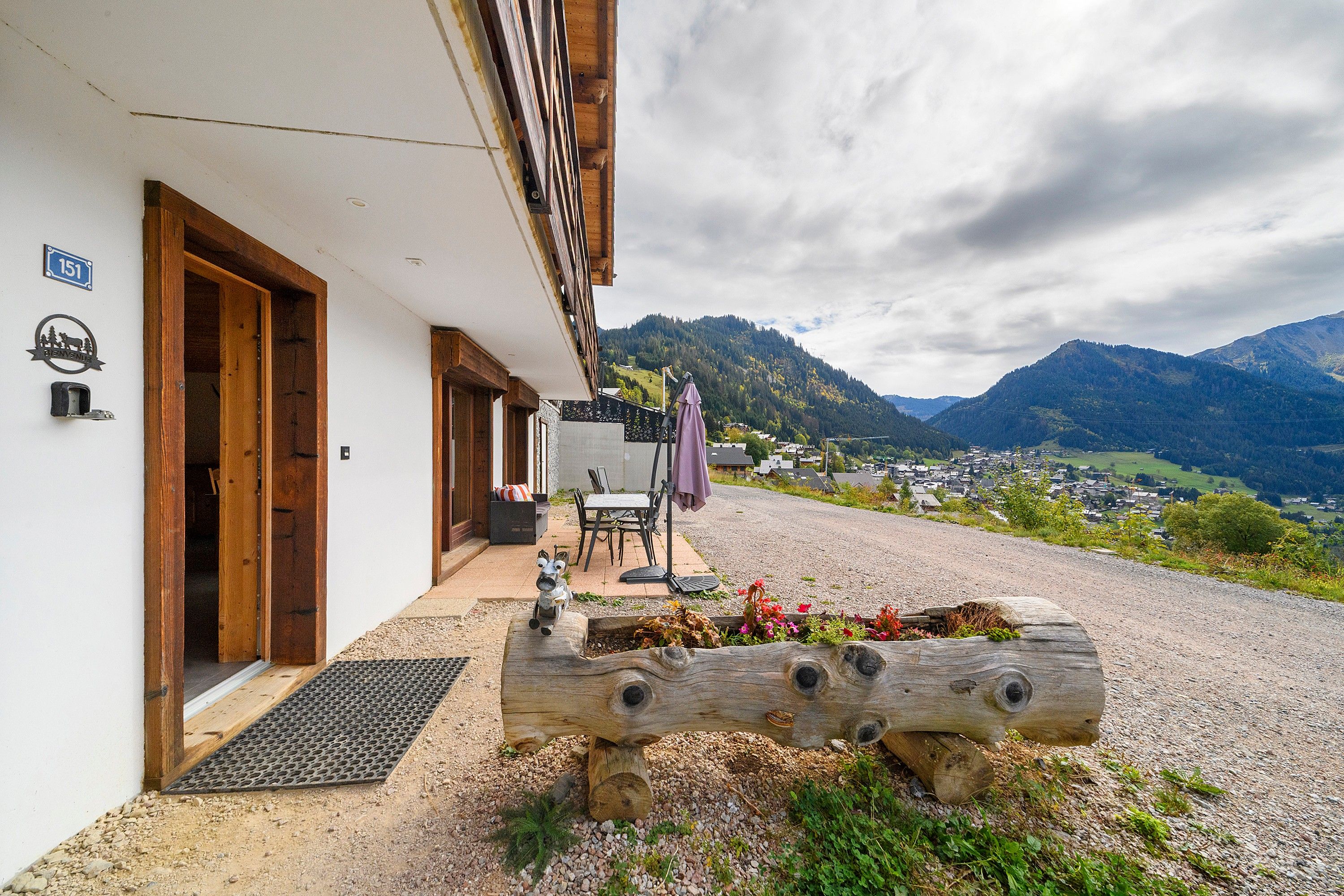 A building with a wooden door and a number sign, a bench with a log seat, and a view of a valley with mountains in the background.