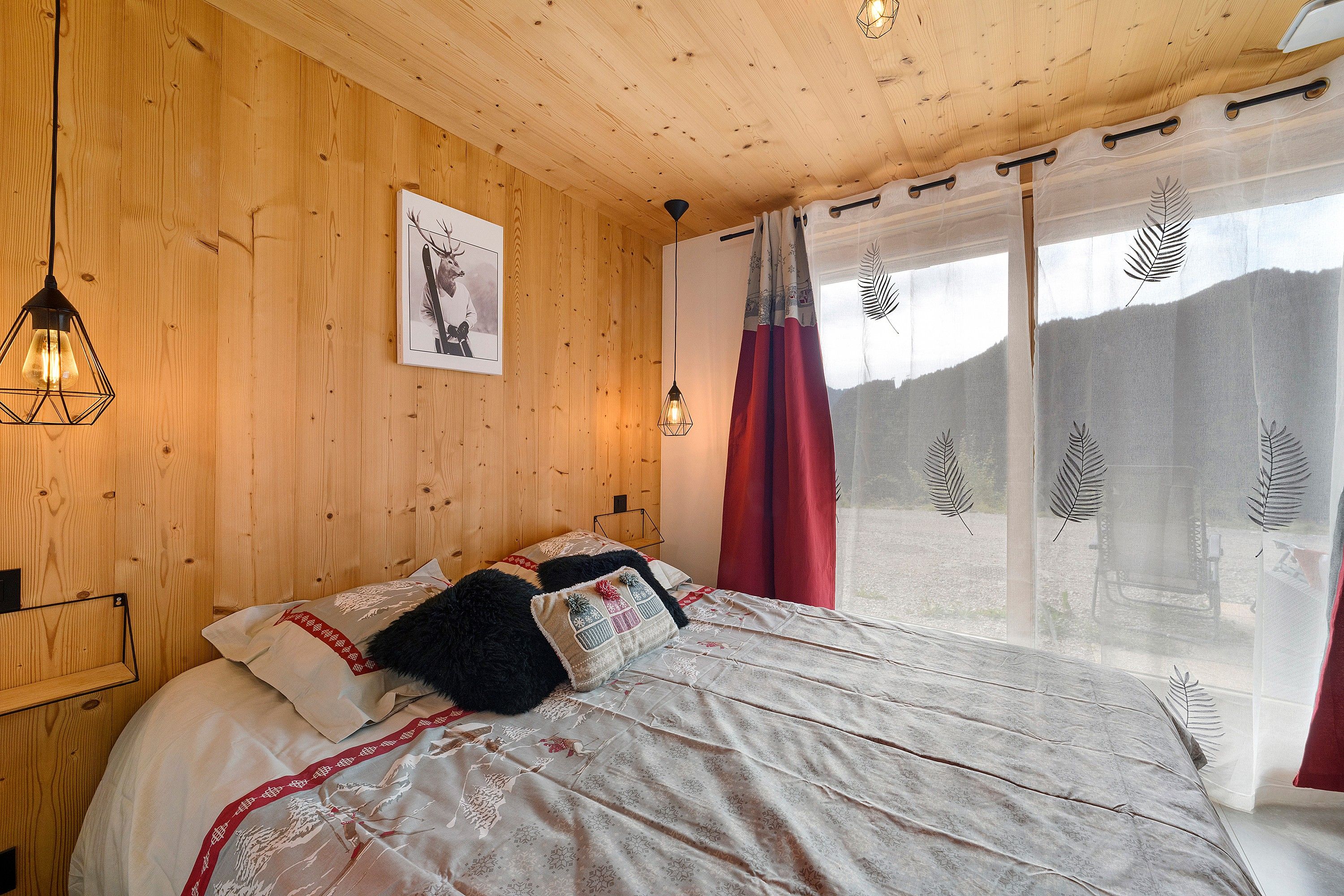 A wooden-paneled room with a bed, a framed picture on the wall, and a window with sheer curtains.