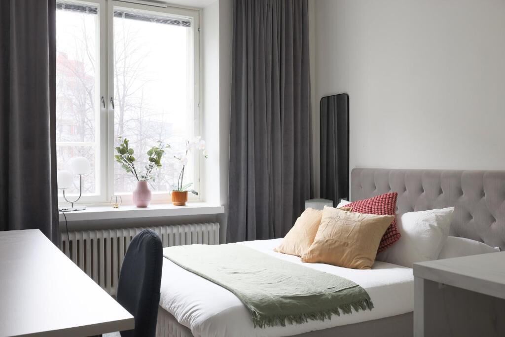 A bedroom with a bed, a bedside table, a chair, a window with curtains, and a radiator.