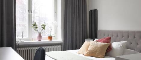 A bedroom with a bed, a bedside table, a chair, a window with curtains, and a radiator.