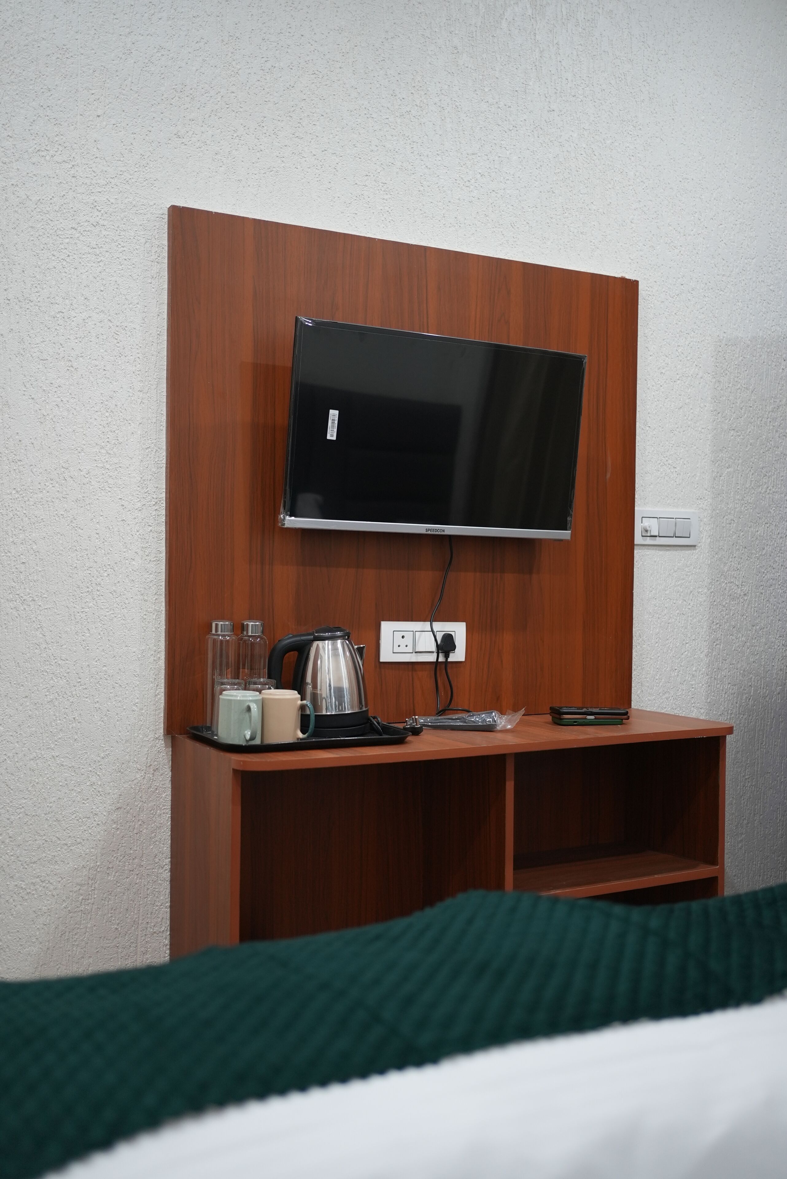 A hotel room with a flat-screen TV mounted on a wooden panel, a small table with a kettle, cups, and a remote control.