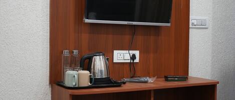 A hotel room with a flat-screen TV mounted on a wooden panel, a small table with a kettle, cups, and a remote control.
