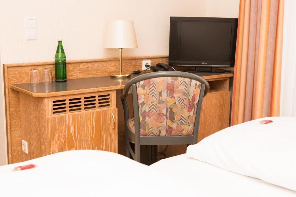 A hotel room with a wooden desk, a television, a patterned chair, and a bed with white bedding.