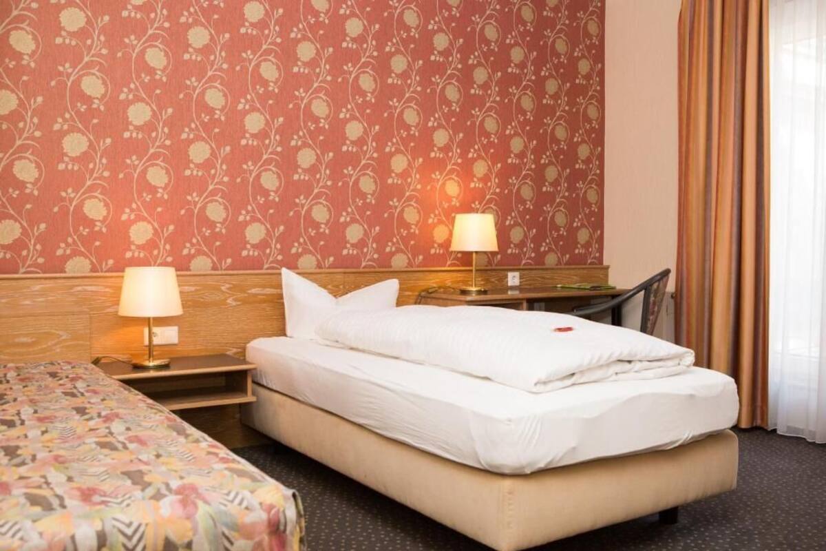 Habitación de hotel con cama, mesitas de noche, lámparas, cabecero de madera y papel tapiz estampado.