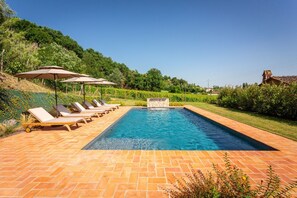 Une piscine rectangulaire, bordée d’une terrasse carrelée, entourée de chaises longues et de parasols, le tout se fondant dans un décor de verdure et de collines.