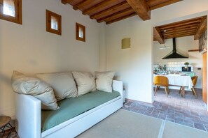 Ein weißes Sofa mit einem grünen Kissen in einem Zimmer mit Fliesenboden, Holzbalken an der Decke und einem durchgehenden offenen Durchgang zur Küche.