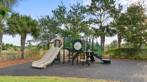 A playground with a slide and climbing structures, surrounded by trees and a fenced area. - Beach House - Salt and Pine at Windmark Beach (Port St. Joe)