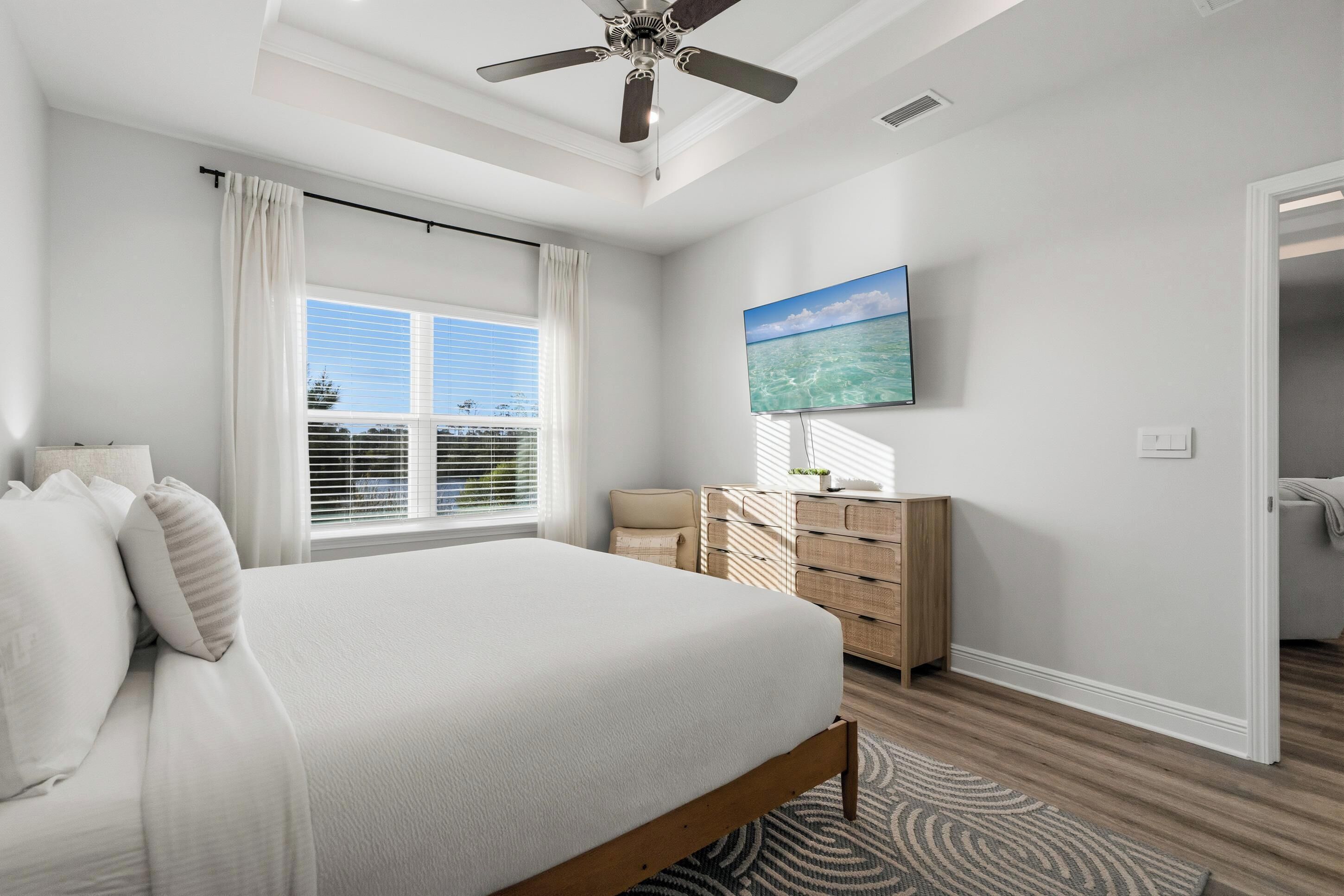 Ein Schlafzimmer mit Bett, Deckenventilator, Kommode, Fernseher und einem Fenster mit Jalousien.