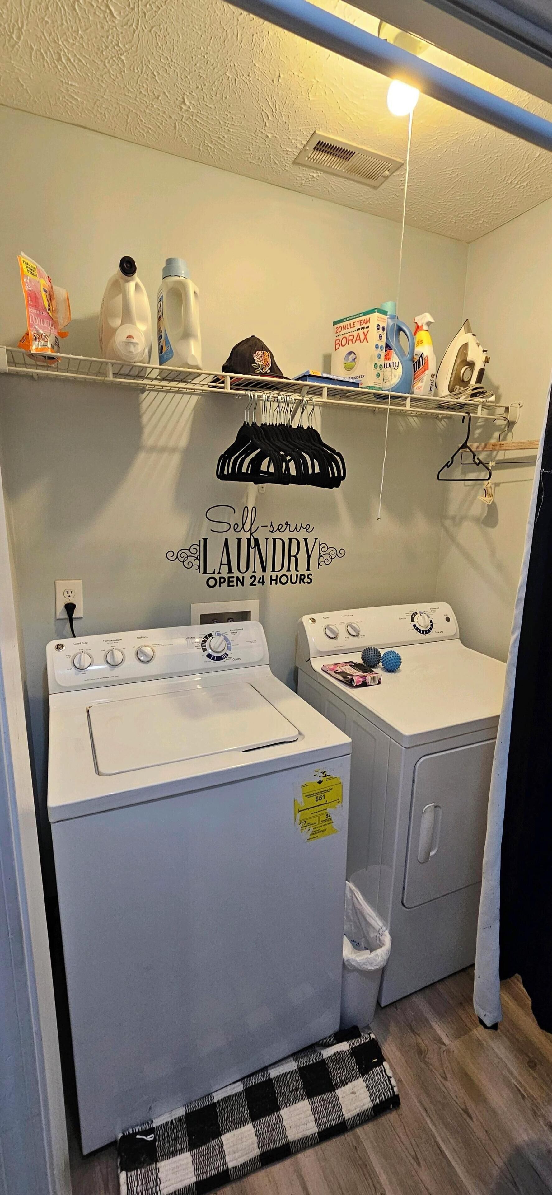 A laundry room with a wall-mounted shelf holding cleaning supplies, a wall-mounted drying rack, a white washing machine, a dryer, and a black and white checkered rug on the floor.