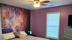Une chambre à coucher comprenant un lit, un ventilateur de plafond, une fenêtre, une commode et une fresque murale sur le thème de la galaxie.