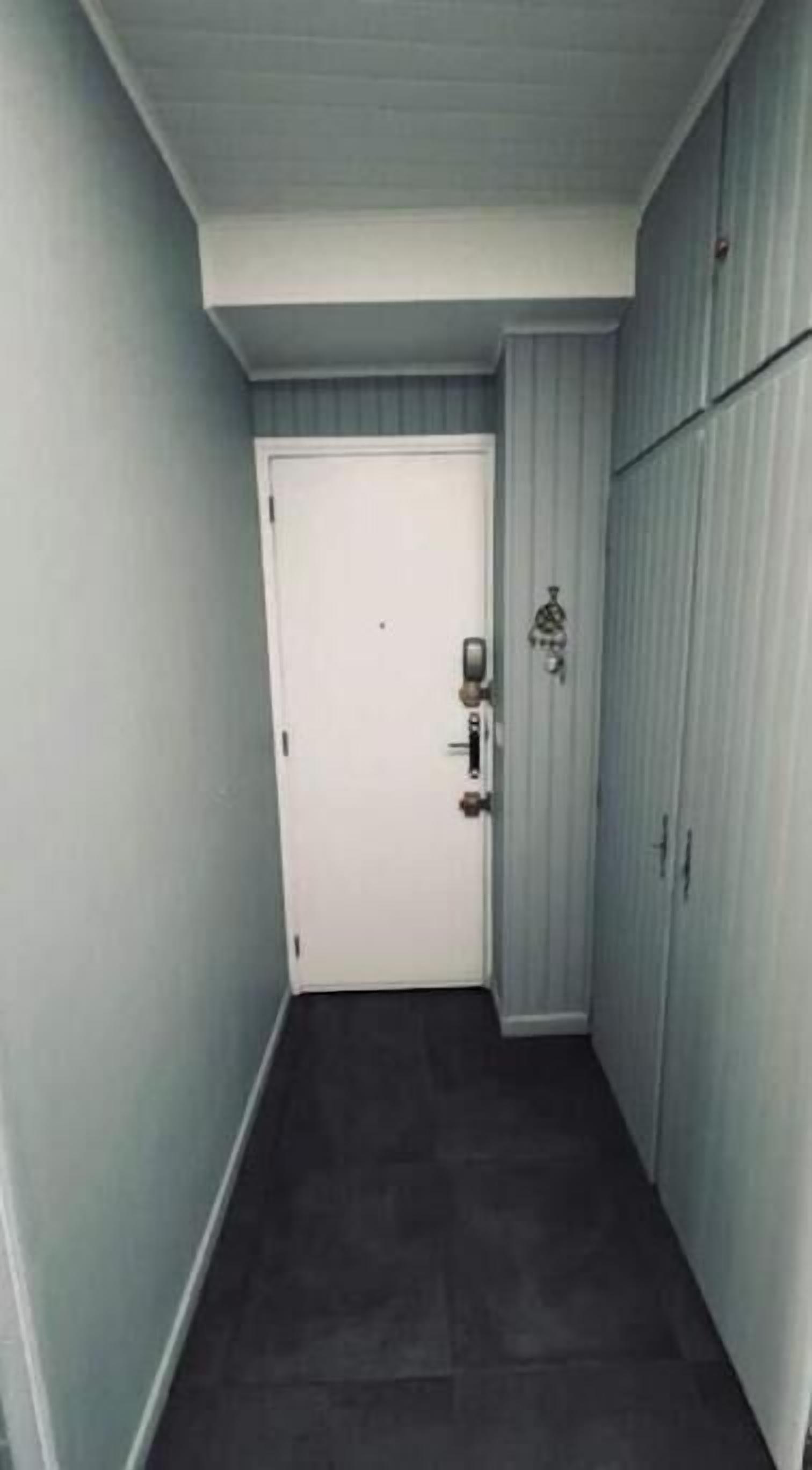 Ein schmaler Flur mit einer weißen Tür, einem Klingel und einem kleinen dekorativen Gegenstand an der Wand.