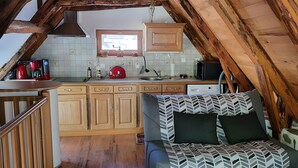 A loft-style bedroom with a bed, a kitchenette, wooden beams, and a window. - Charming Small Cottage for 2/3 Guests – La Chèvrerie (Saint-Vincent-de-Salers)