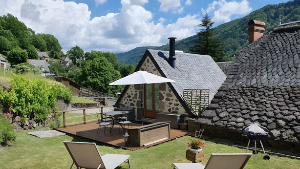 A cozy outdoor seating area with a table and chairs, a stone house with a sloped roof, and a view of a lush green valley. - Charming Small Cottage for 2/3 Guests – La Chèvrerie (Saint-Vincent-de-Salers)