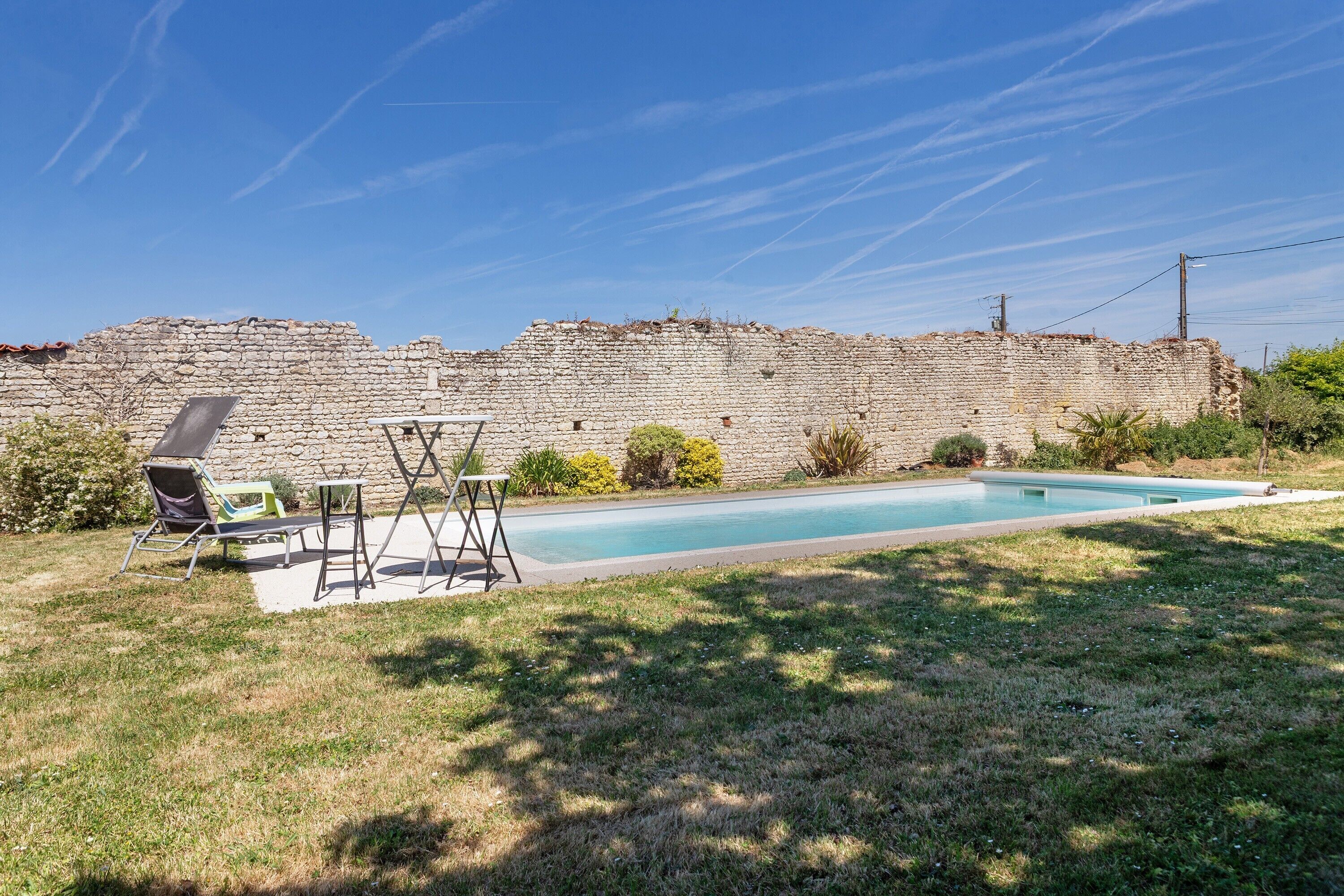 Une piscine avec un mur de pierre en arrière-plan, une pelouse et du mobilier d’extérieur.