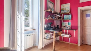 Ein Zimmer mit Schreibtisch, Regalen mit diversen Gegenständen, einem Fenster und einer Tür.