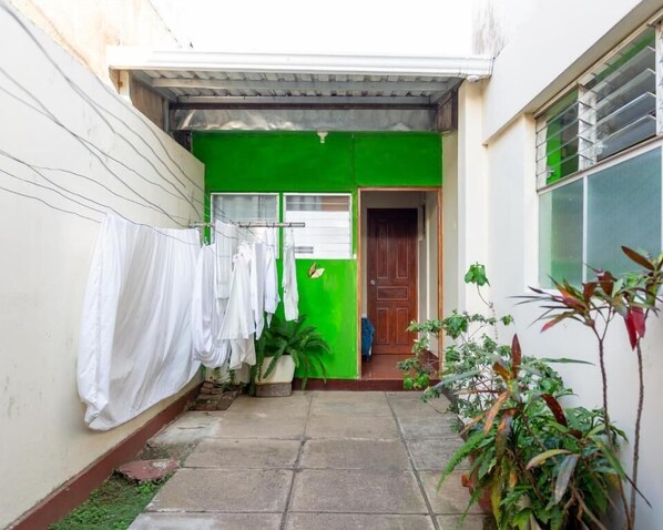 A narrow courtyard with a green wall, a clothesline with white garments, a door, and potted plants.
