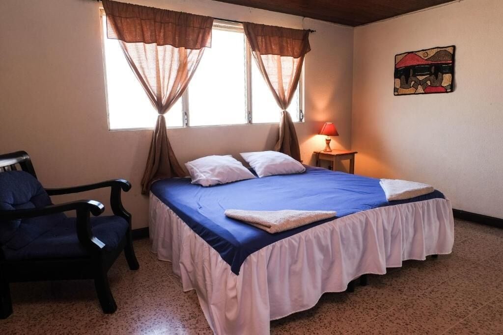 A bedroom with a bed, a chair, a window with curtains, a nightstand with a lamp, and a wall art piece.