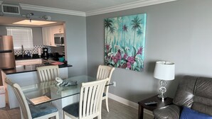 Un comedor con mesa de vidrio, sillas blancas, una cocina al fondo y un cuadro grande con motivos florales en la pared.