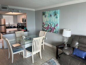Een eetgedeelte met een glazen tafel, witte stoelen, een keuken op de achtergrond en een groot bloemschilderij aan de muur.