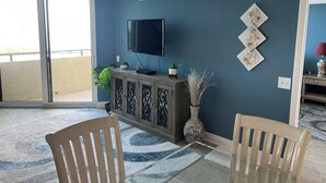 Una sala de estar con televisor, aparador, dos sillas, una planta grande y un adorno de pared.