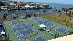Una cancha de tenis con red, rodeada por un estacionamiento y un puerto deportivo con barcos.