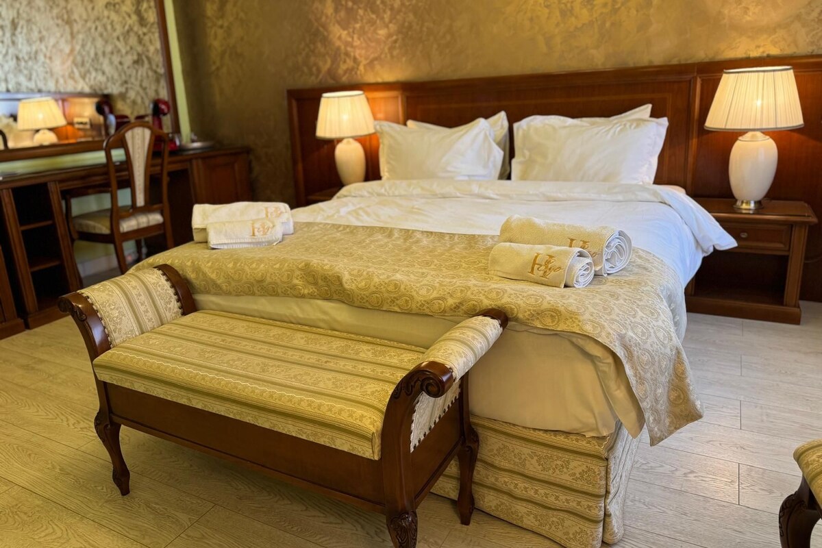 A hotel room with a large bed, two bedside tables with lamps, a wooden bench, and a desk with a chair.