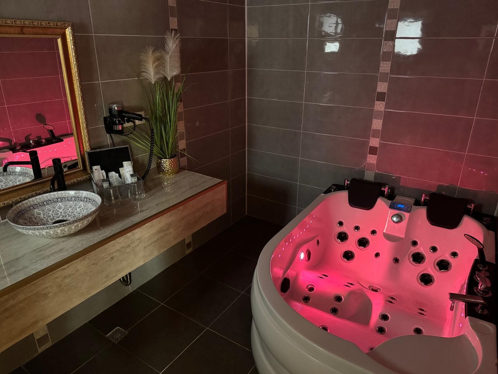 A bathroom with a vanity, a large mirror, a bowl sink, a potted plant, and a spa tub with pink lighting.