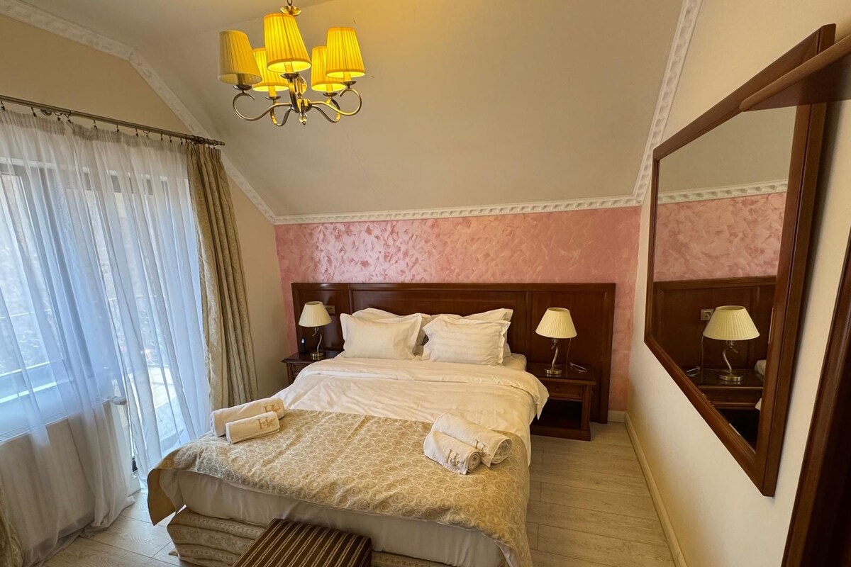 A hotel room with a large bed, two bedside tables with lamps, a large mirror, and a window with sheer curtains.