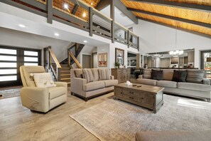 A spacious living room with a high ceiling, a staircase, and a kitchen in the background. - Evergreen (Big Bear Lake)