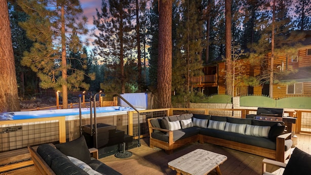 A wooden deck with a hot tub, a wooden sofa, and a grill area surrounded by tall trees. - Evergreen (Big Bear Lake)