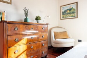 A wooden dresser with multiple drawers, a small potted plant, and a lamp on a bedside table. A white chair with a cushion is positioned against the wall. - GuestHost - Enchanting villa located within an agricultural estate on the Tuscan hills, surrounded by nature and with a breathtaking view of the surrounding countryside. The property, equipped with private outdoor parking, outdoor swimming pool, gym area (Palaia)