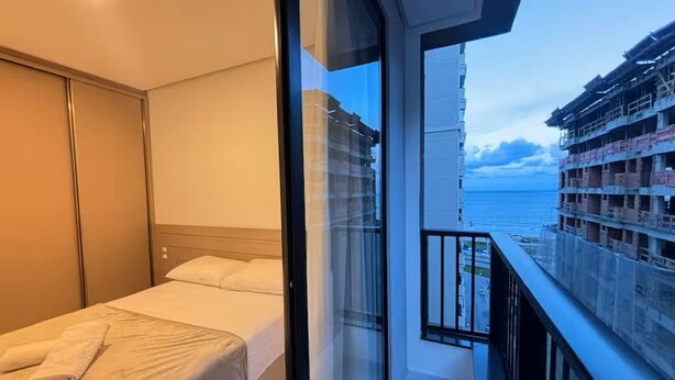 A bedroom with a bed, a nightstand, and a window with a view of a building and a body of water. - Ocean View, New Apartment with Garage and BBQ (Itapema)