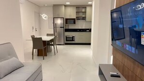 A modern living room with a sofa, a dining area with chairs, a kitchen with cabinets and appliances, and a TV mounted on the wall. - Ocean View, New Apartment with Garage and BBQ (Itapema)