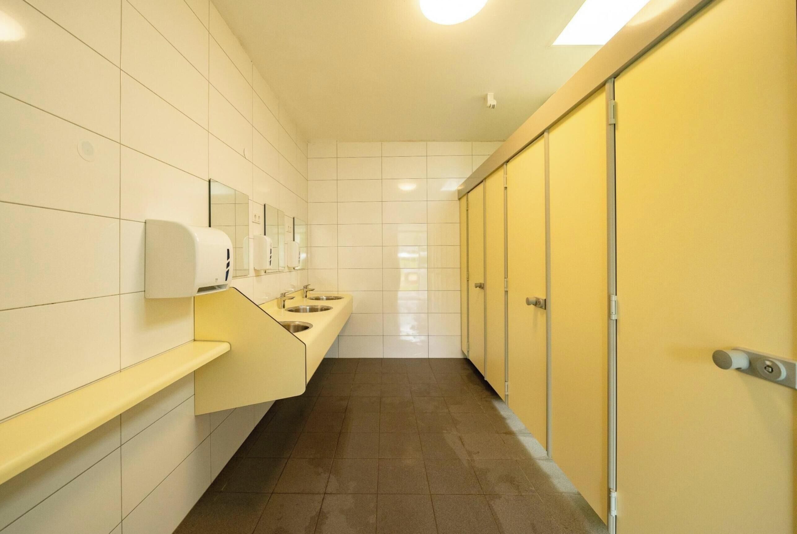 A public restroom with white tiled walls, a yellow sink area, and a row of yellow doors.