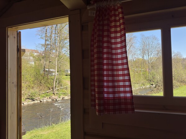 A room with a window overlooking a river, a red and white checkered curtain, and a view of trees and buildings. - Eifelhutte auf dem Campingpark (Waxweiler)