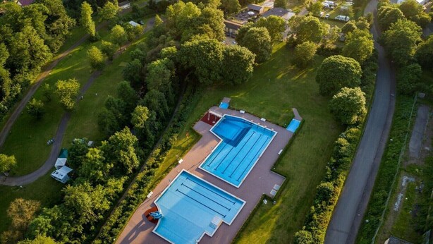 An aerial view of a campground with multiple RV sites, a swimming pool, and surrounding forested hills. - Eifelhutte auf dem Campingpark (Waxweiler)