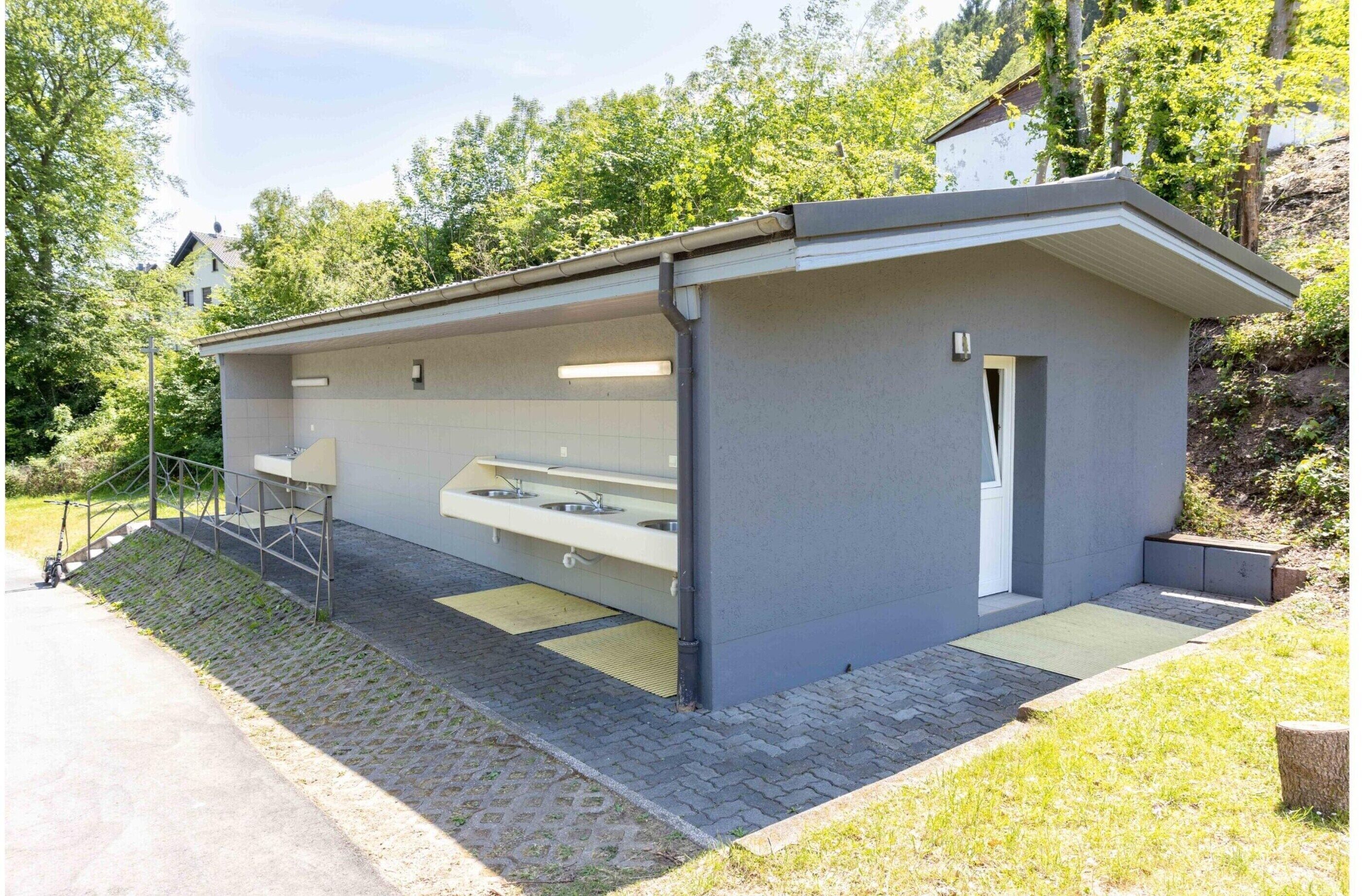A modern outdoor restroom with a large sink and a door, surrounded by a paved area and greenery.