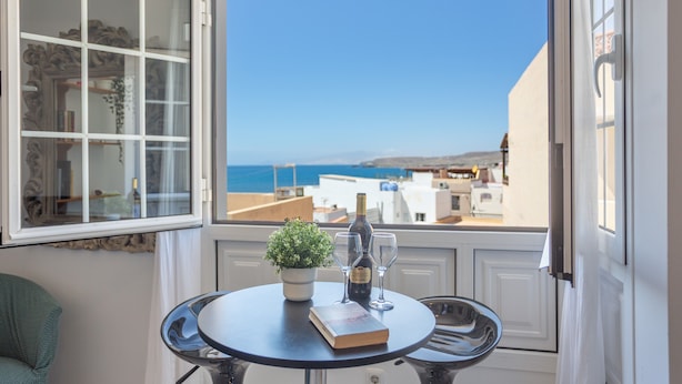 Una habitación con vistas al mar, una mesa con un libro y vino, y una planta en maceta.