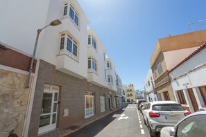 Une rue étroite bordée d’immeubles à plusieurs étages, de voitures garées et d’un ciel dégagé.